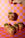 A hand holds the OCTAEVO Pistachio Cruffin Ornament above four others on a pink and orange checkered surface.