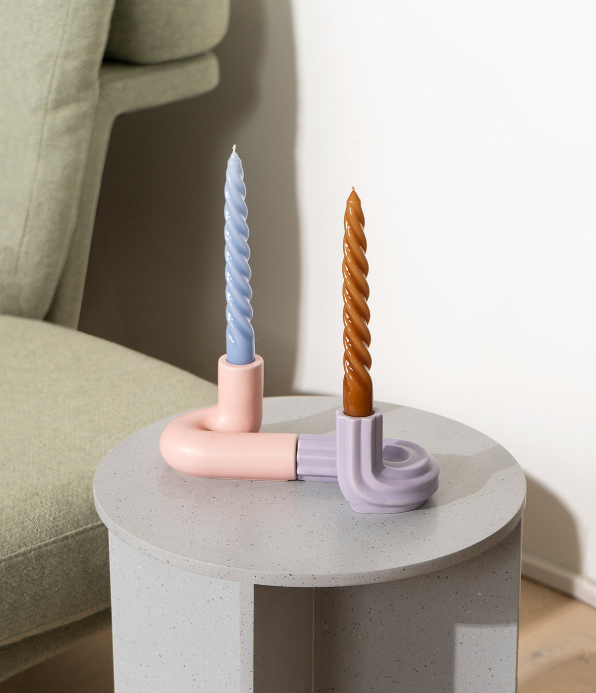 Two twisted candles, one blue and one brown, in pastel pink and purple holders on a round gray side table.