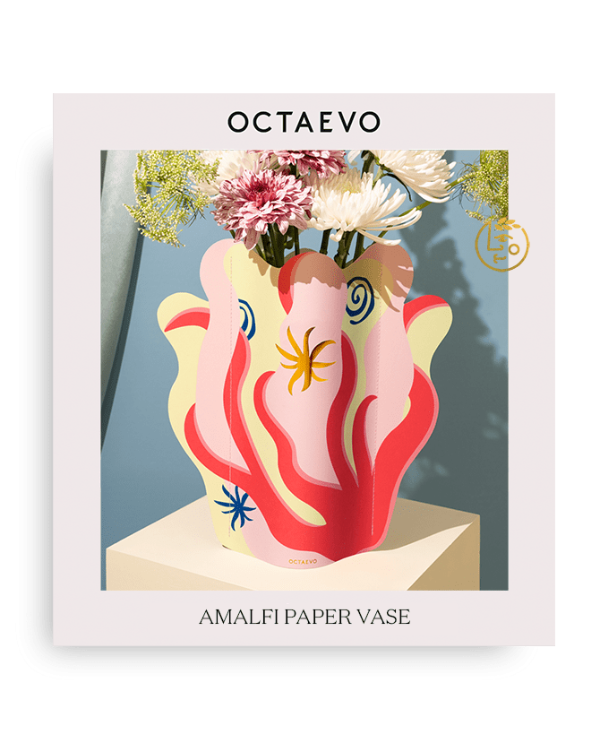 Le vase en papier Amalfi d'Octaevo présente des motifs éclatants et accueille des fleurs blanches et roses, sur une table beige.