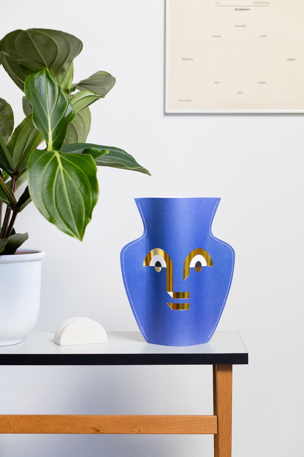 The OCTAEVO Apollo Paper Vase with a face design sits on a table beside a potted plant and white decor.