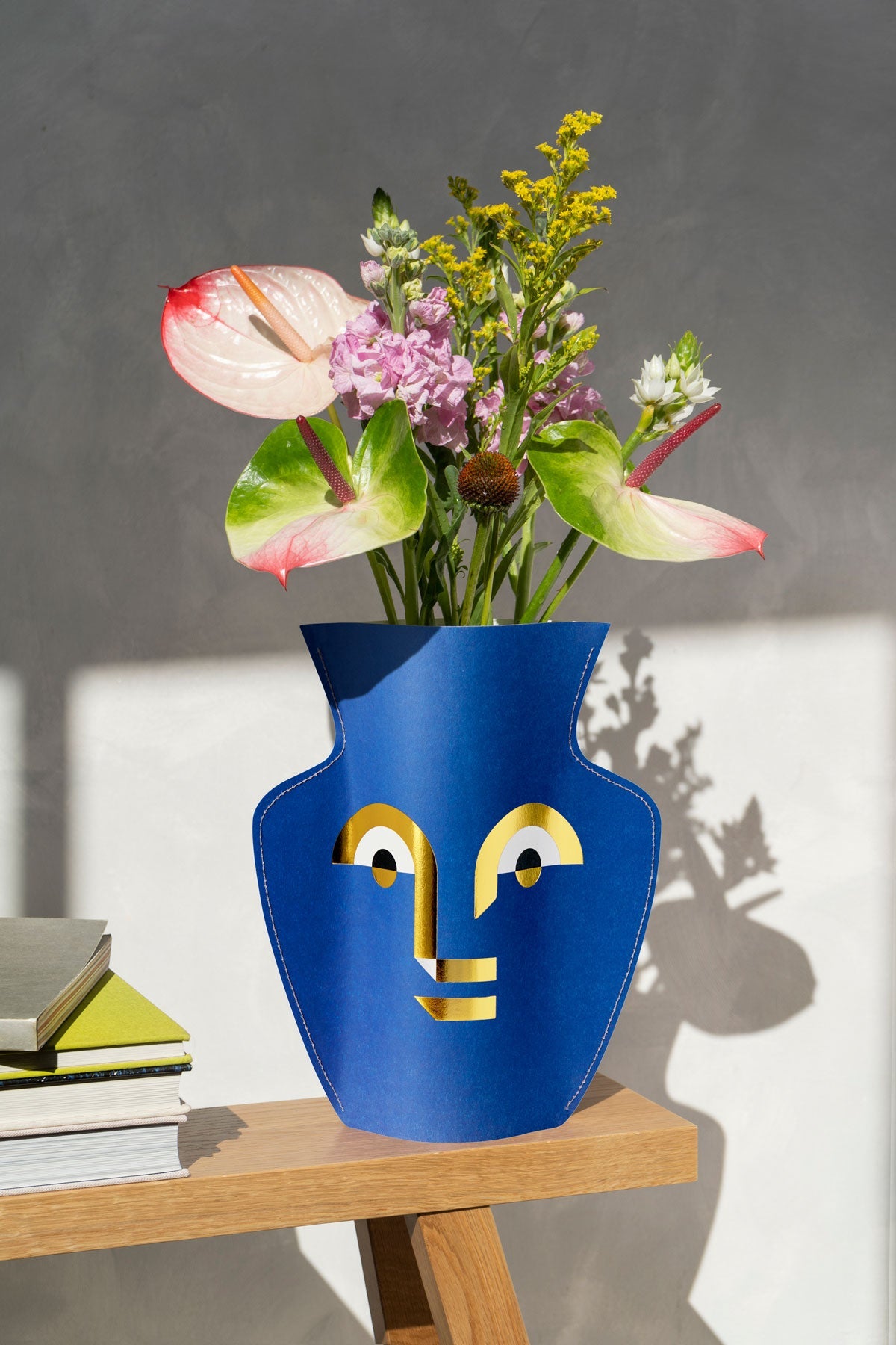 An OCTAEVO Apollo Paper Vase with colorful flowers sits on a wooden table beside stacked books in sunlight.