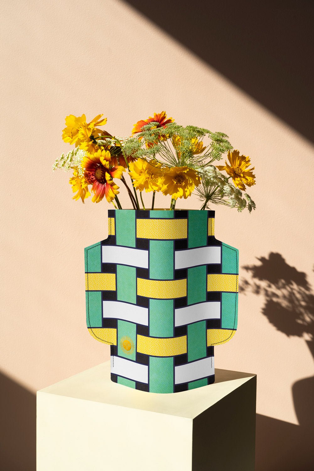 Yellow flowers in the OCTAEVO Artesania Paper Vase with teal, yellow, and black patterns on a white pedestal in sunlight.
