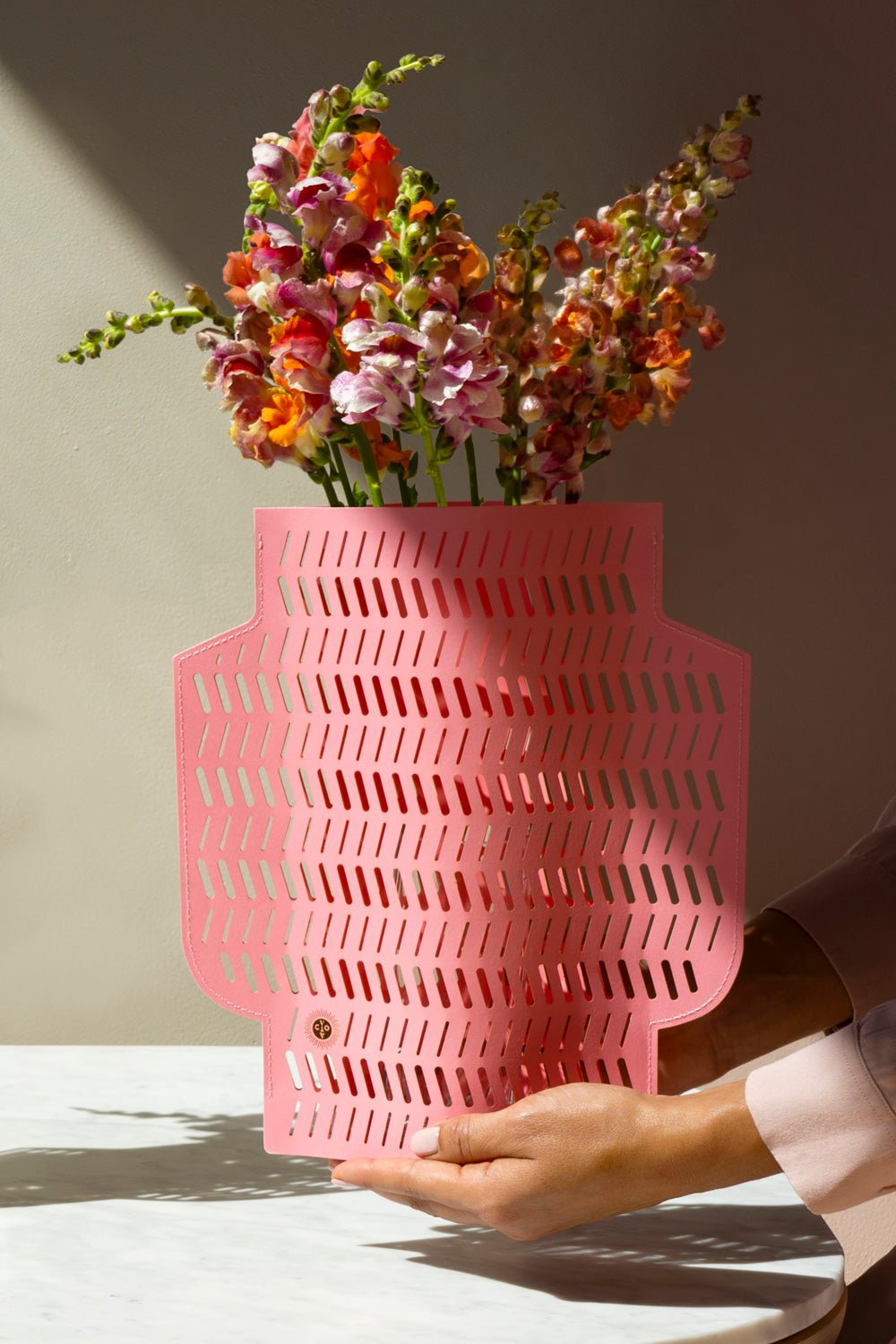 A hand holds the Aurea Paper Vase by OCTAEVO, filled with orange and pink flowers, casting patterned shadows.