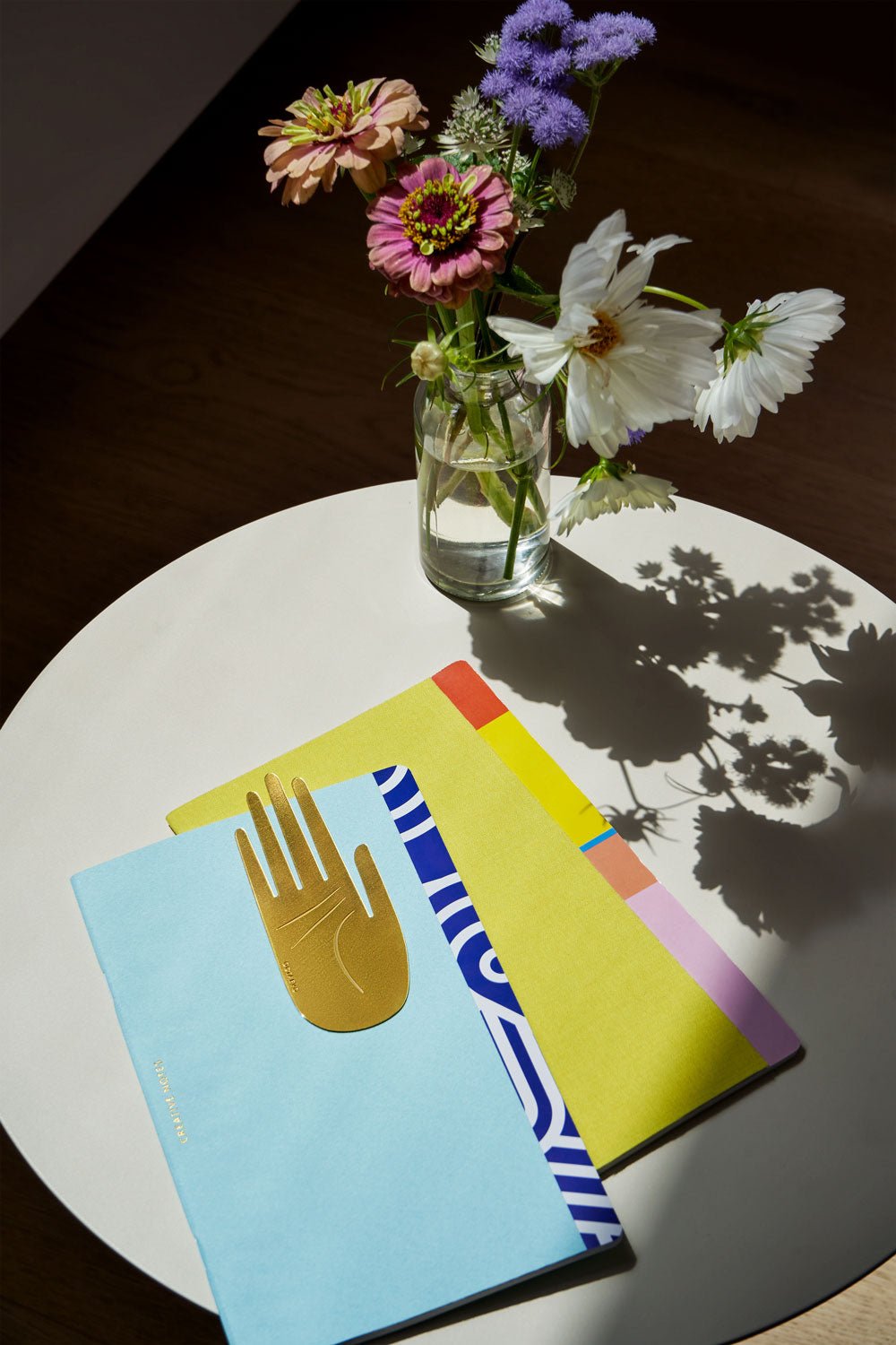 Greco Creative Notes by OCTAEVO and vibrant flowers rest on a sunlit round white table with two cheerful friends.