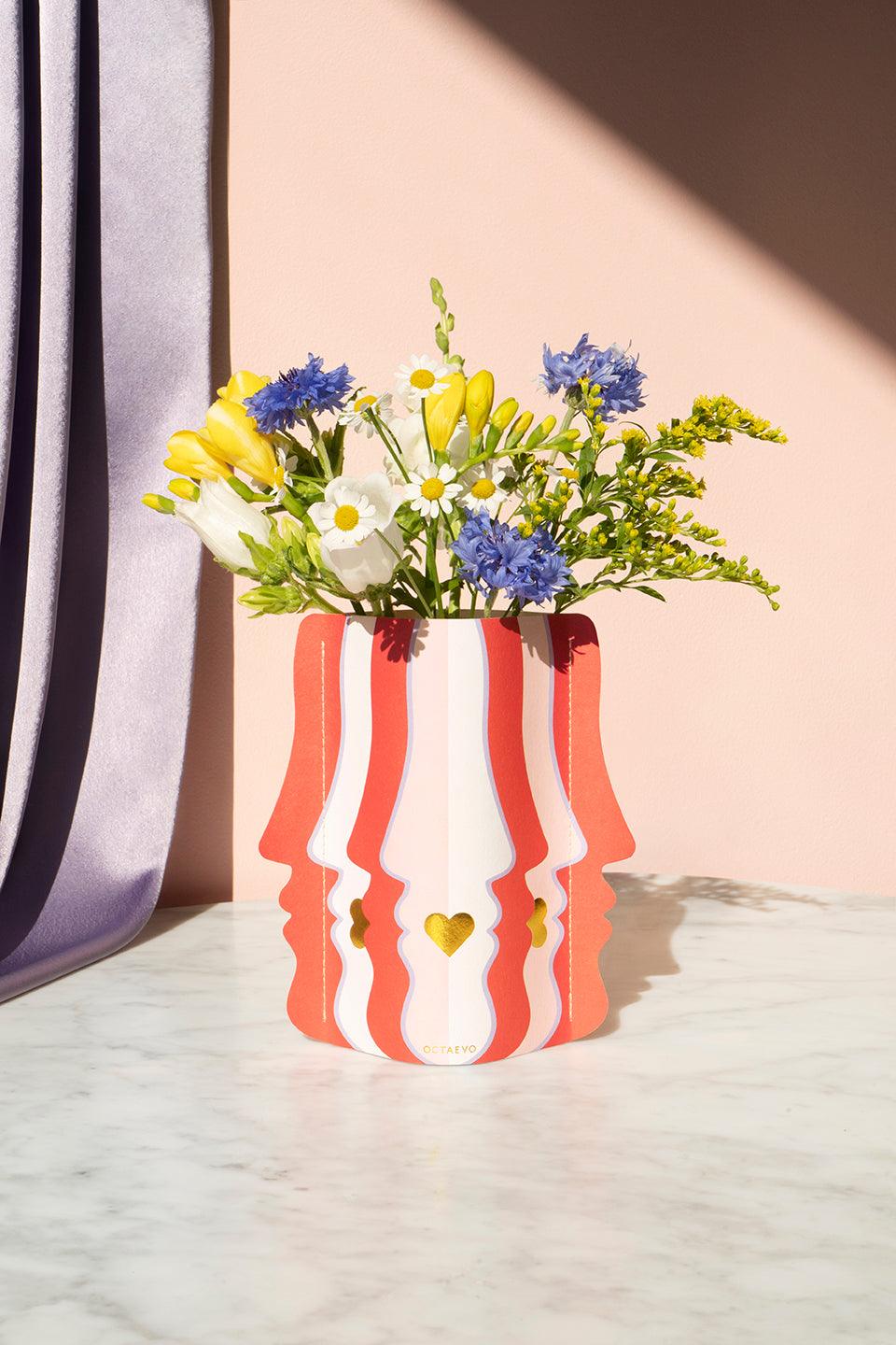 The Octaevo Beso Mini Paper Vase with face profiles holds flowers—an original gift idea for any decor.