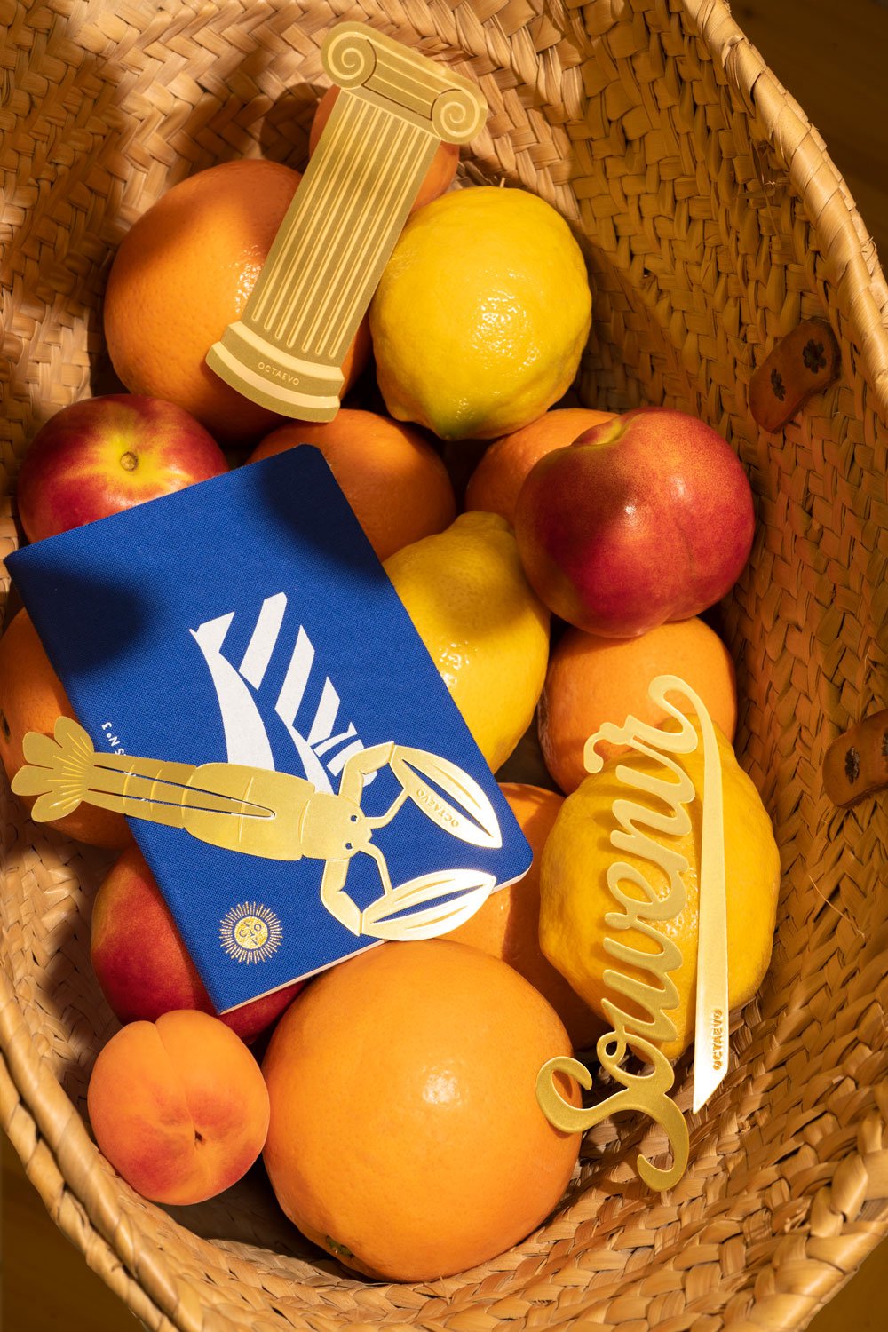 A woven fruit basket, passport notebook, OCTAEVO Bookmark Artemis (gold), Bon Vivant gold and Souvenir gold.