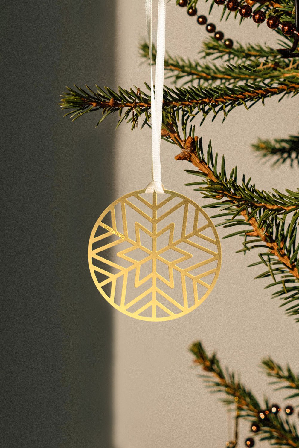 A green Christmas Bauble Bookmark by OCTAEVO hangs from a branch with a white ribbon, glowing in warm sunlight.