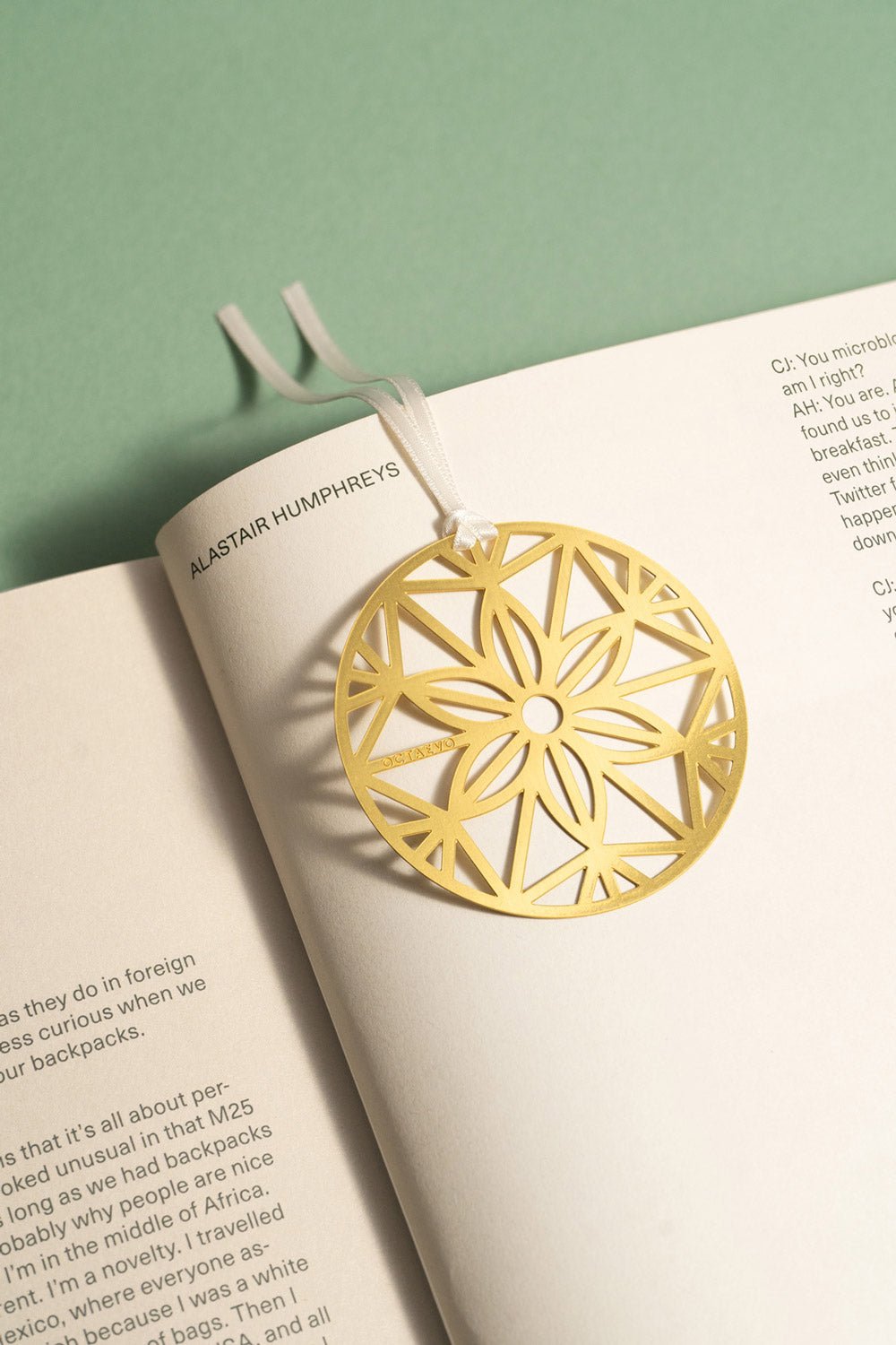 The OCTAEVO Bookmark Christmas Bauble, gold and floral-patterned, lies on an open book atop a green surface.