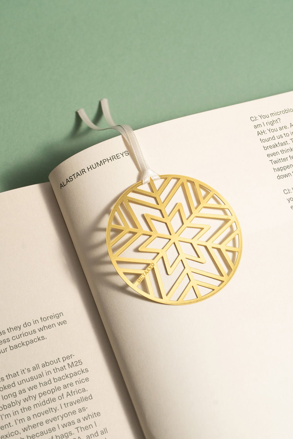 The OCTAEVO Bookmark Christmas Bauble—a gold geometric snowflake with a white ribbon—adorns your open book festively.