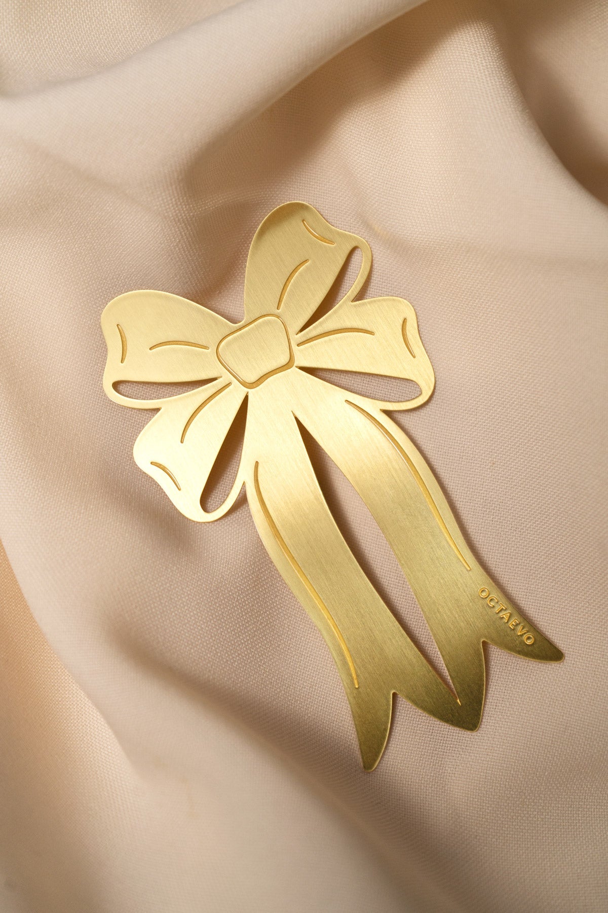 Octaevo’s Bookmark Coquette, a gold ribbon-shaped brooch, rests elegantly on soft light beige fabric.