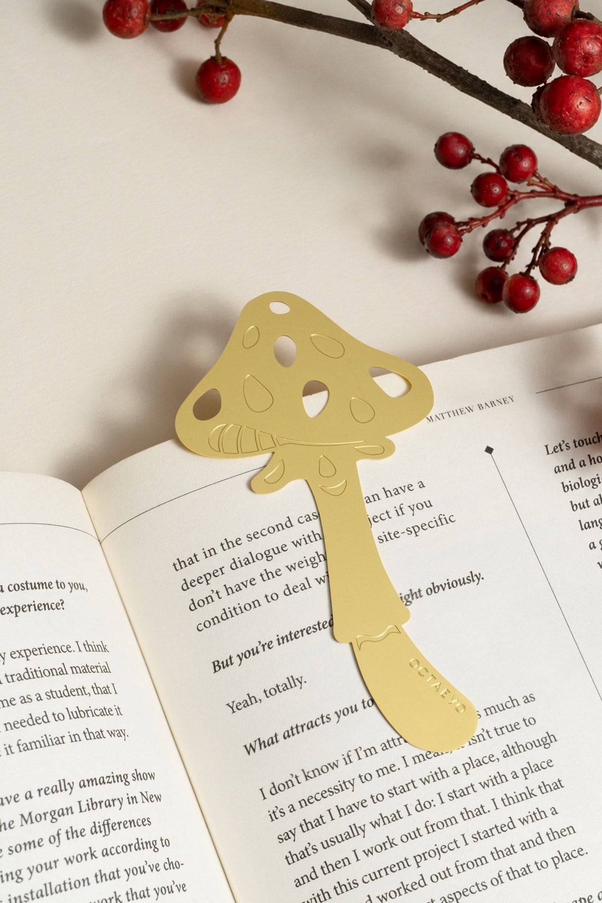 The OCTAEVO Bookmark Magic Mushroom, a gold metal marker, is shown on an open book with red berries nearby.