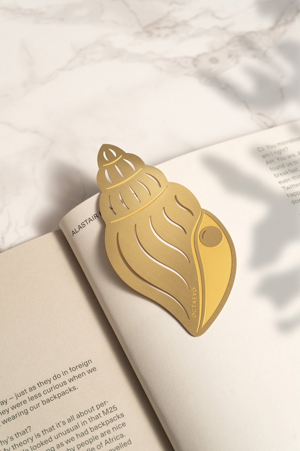 The OCTAEVO Bookmark Pearl Shell rests on the open pages of a book displayed on a marble surface.