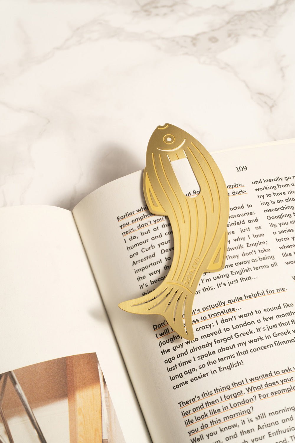 The OCTAEVO Bookmark Sardine, finely cut from brass, rests on the open pages of a book on marble.