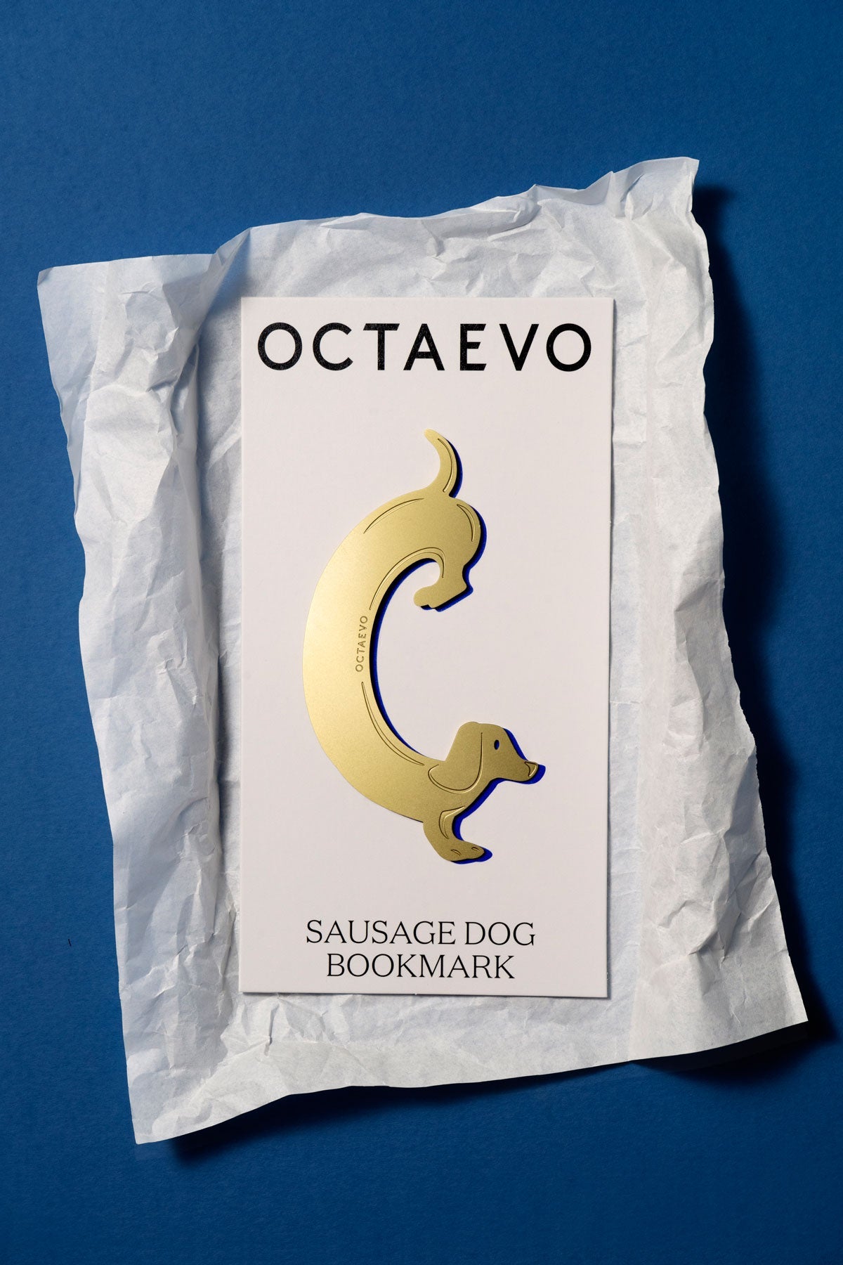 OCTAEVO’s Bookmark Sausage Dog in gold brass sits atop white paper—a fun reading companion on a blue background.