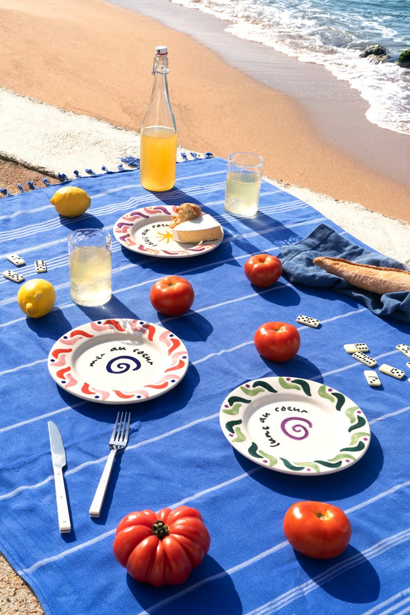 Ein Picknick auf einer blauen Decke mit OCTAEVO Cala Secreta Tellern, Tomaten, Zitronen, Getränken, Brot und Dominosteinen.