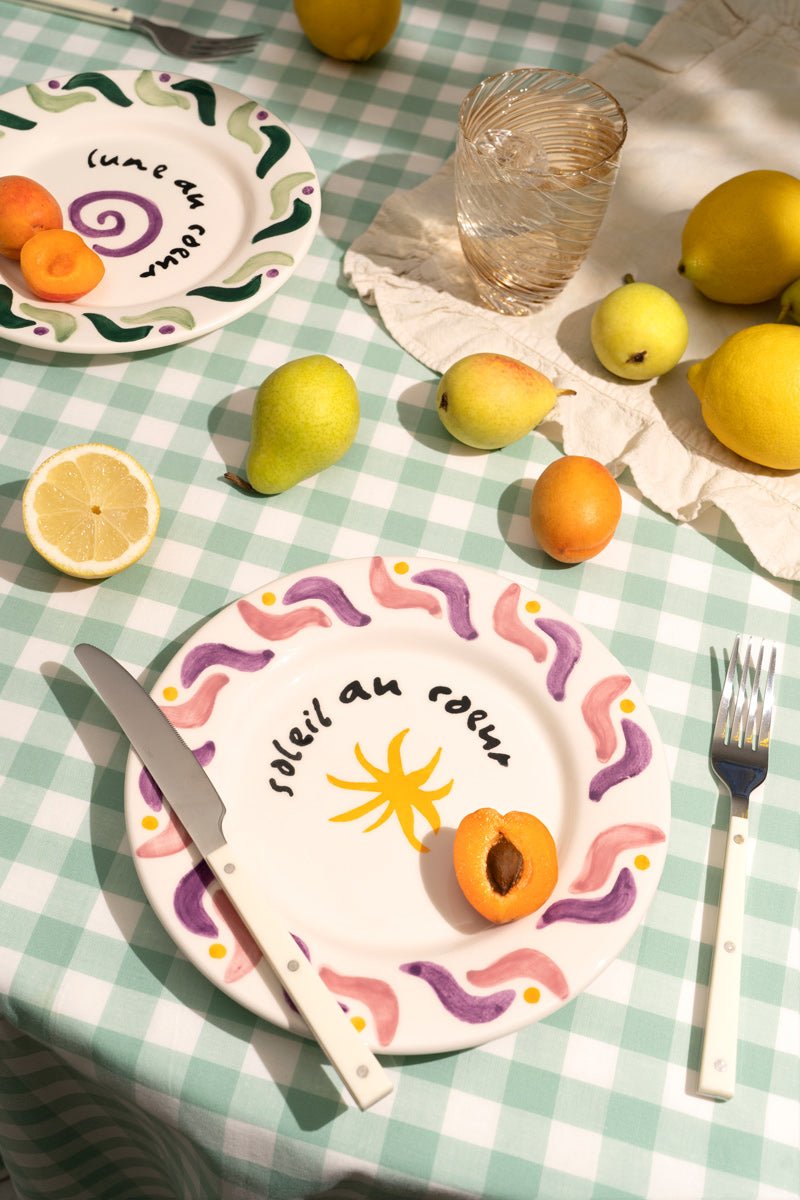 A colorful table with fruit, cutlery, and Octaevo’s hand-painted Cala Secreta Plate on a checkered cloth.