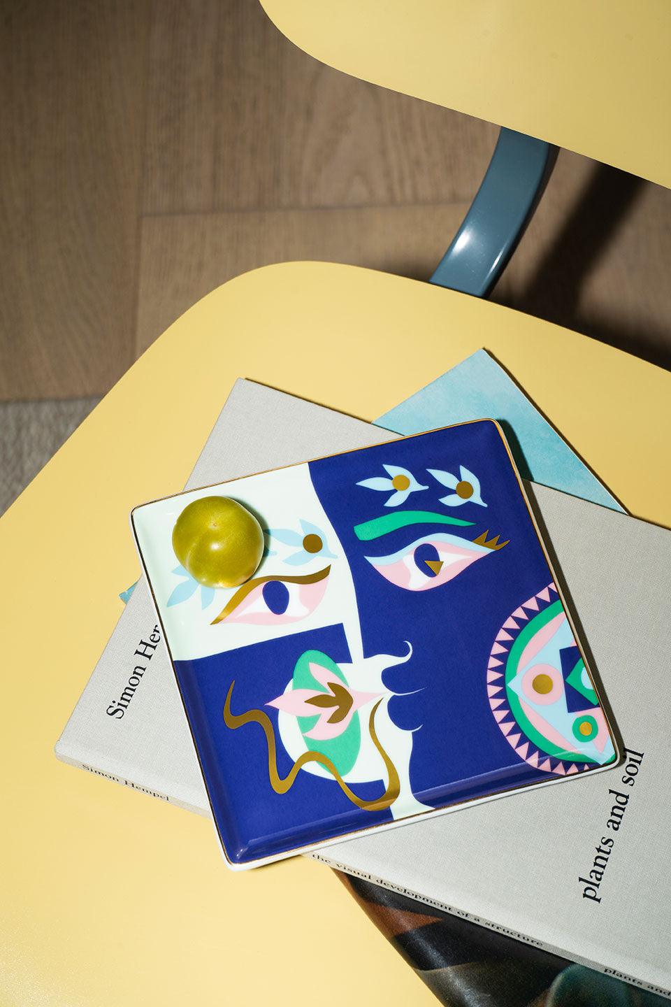 A lime on an OCTAEVO Claire Johnson Ceramic Tray — Blue sits atop books on a yellow chair with a wood floor.