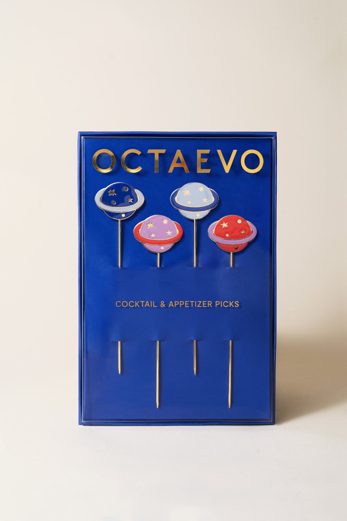 A blue Octaevo box holds four Cosmos gold-plated cocktail picks topped with colorful, planet-shaped designs.