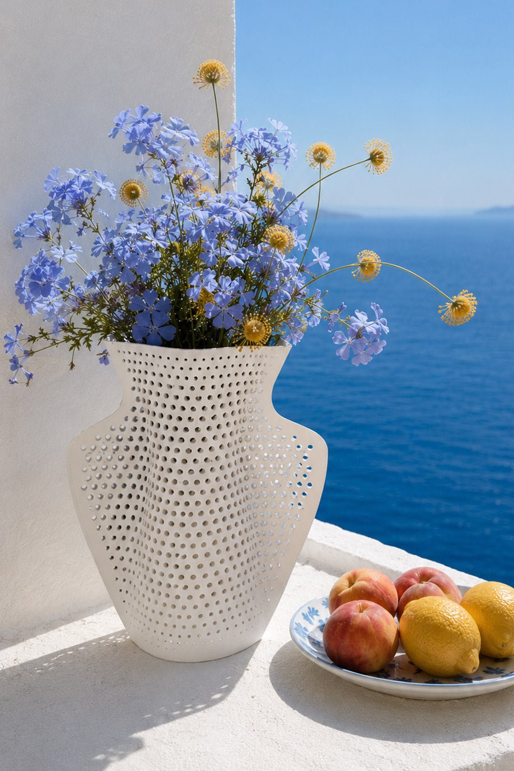 The OCTAEVO Cyano Paper Vase with blue and yellow flowers sits by peaches and lemons on a ledge overlooking the sea.