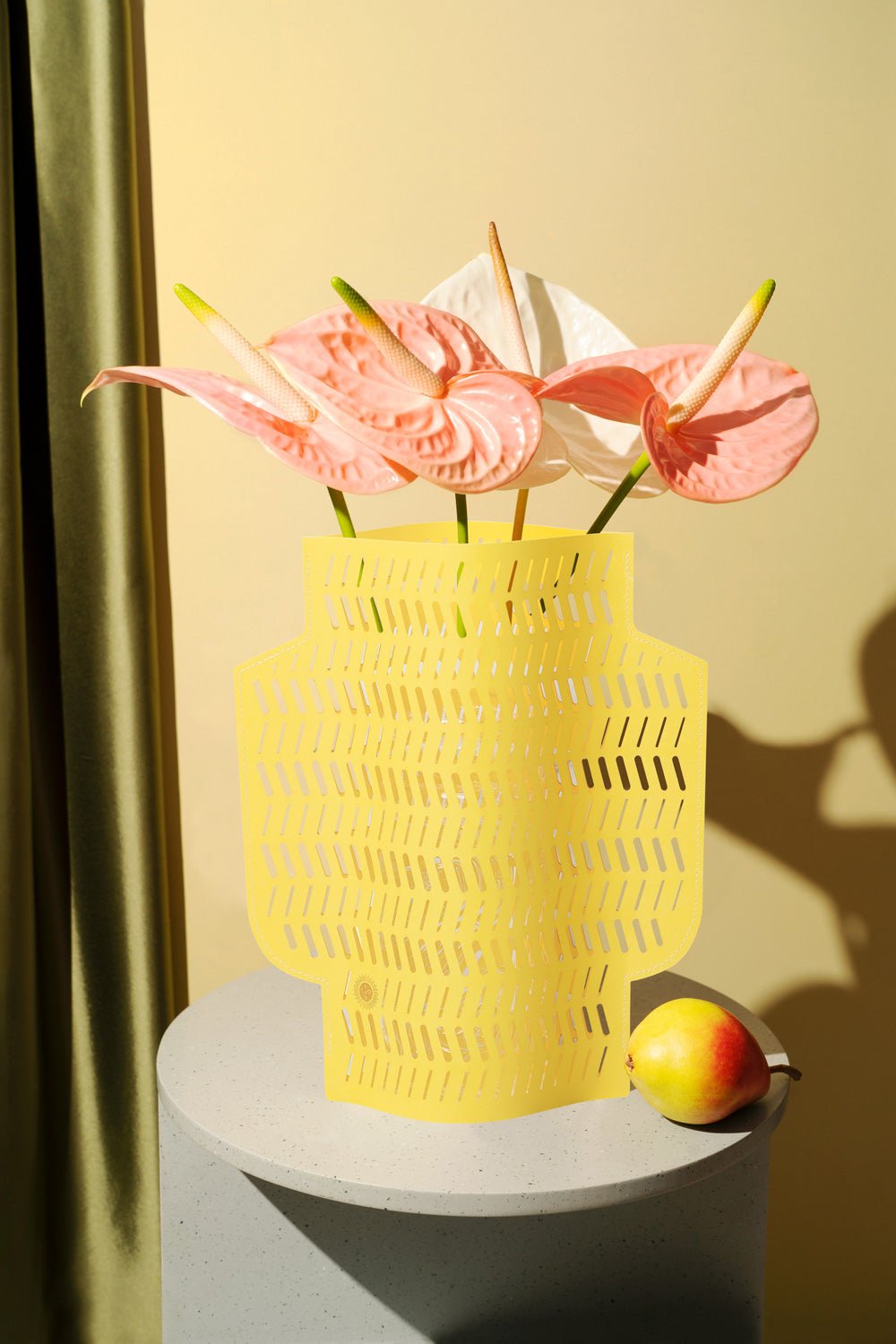 OCTAEVO's Dendra Paper Vase in yellow with pink anthuriums and a pear on a gray pedestal, beige background.