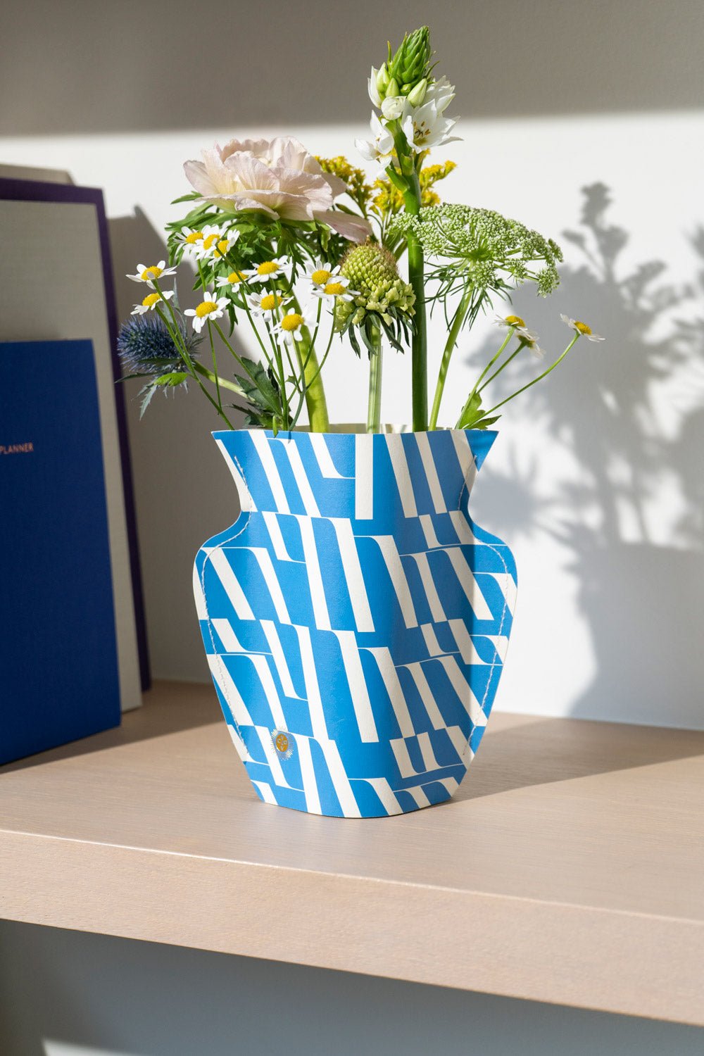 A Domus Mini Paper Vase by OCTAEVO with fresh flowers sits on a light wooden shelf in sunlight.