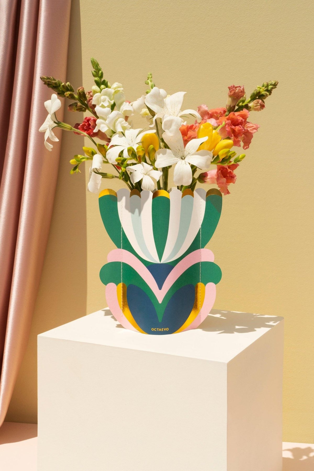 The Elysia Mini Paper Vase by Octaevo with fresh flowers sits on a white pedestal against a pink-curtained beige backdrop.