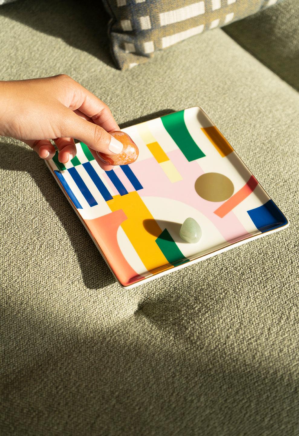 A hand places a stone on the Octaevo Emporio Ceramic Tray, a colorful gift tray resting on a green textured surface.