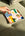 A hand places a stone on the Octaevo Emporio Ceramic Tray, a colorful gift tray resting on a green textured surface.