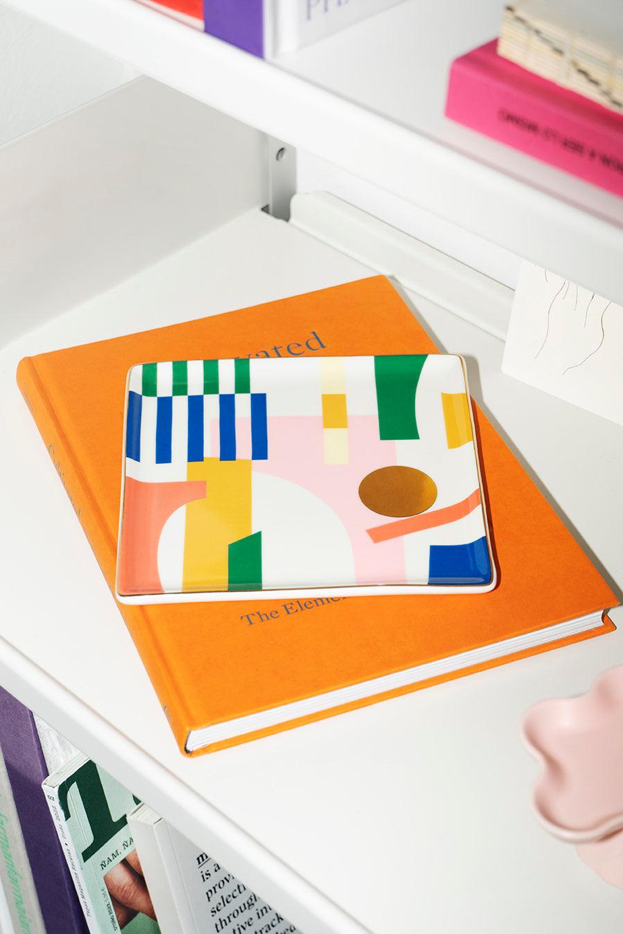 The Emporio Ceramic Tray by Octaevo sits on an orange book, adding a pop of style to a white shelf.