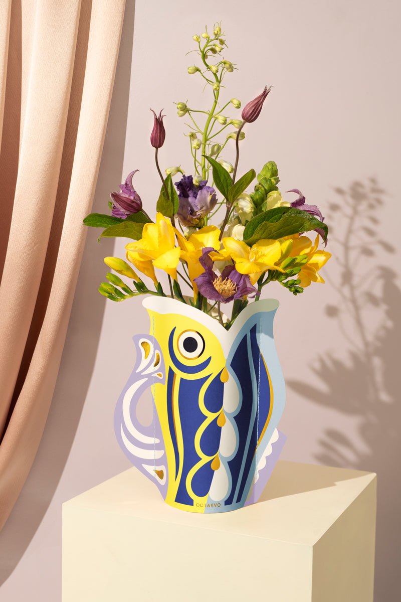 The Fish Mini Paper Vase in purple by OCTAEVO with an abstract bird design holds flowers beside a light pink curtain.