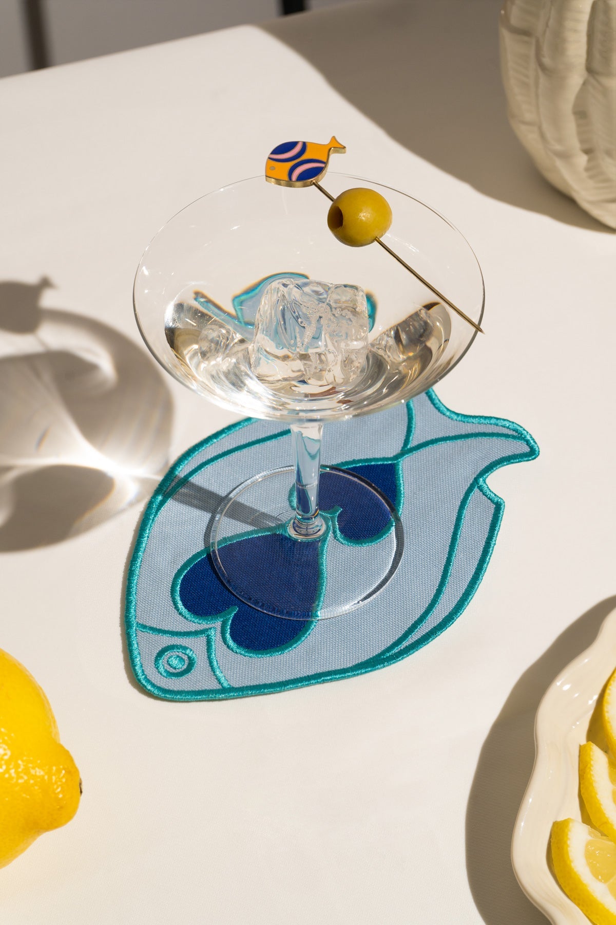 A martini with an olive rests on an Octaevo Fish Splash Coaster in blue, beside a lemon and its fish-shaped shadow.