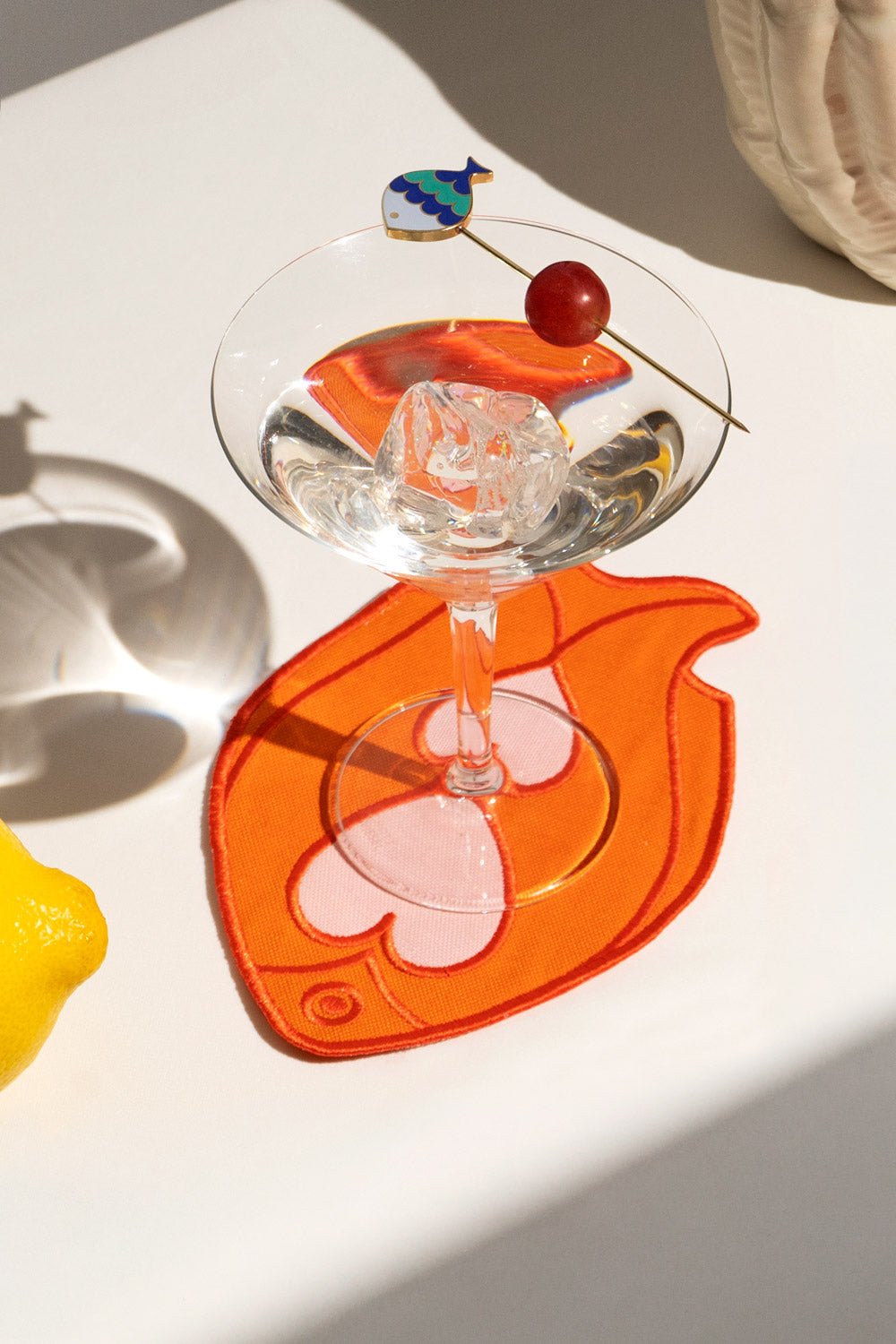 A martini sits on OCTAEVO Fish Splash Coasters (Set of 4) in orange, garnished with grape and fish skewer, beside a lemon.