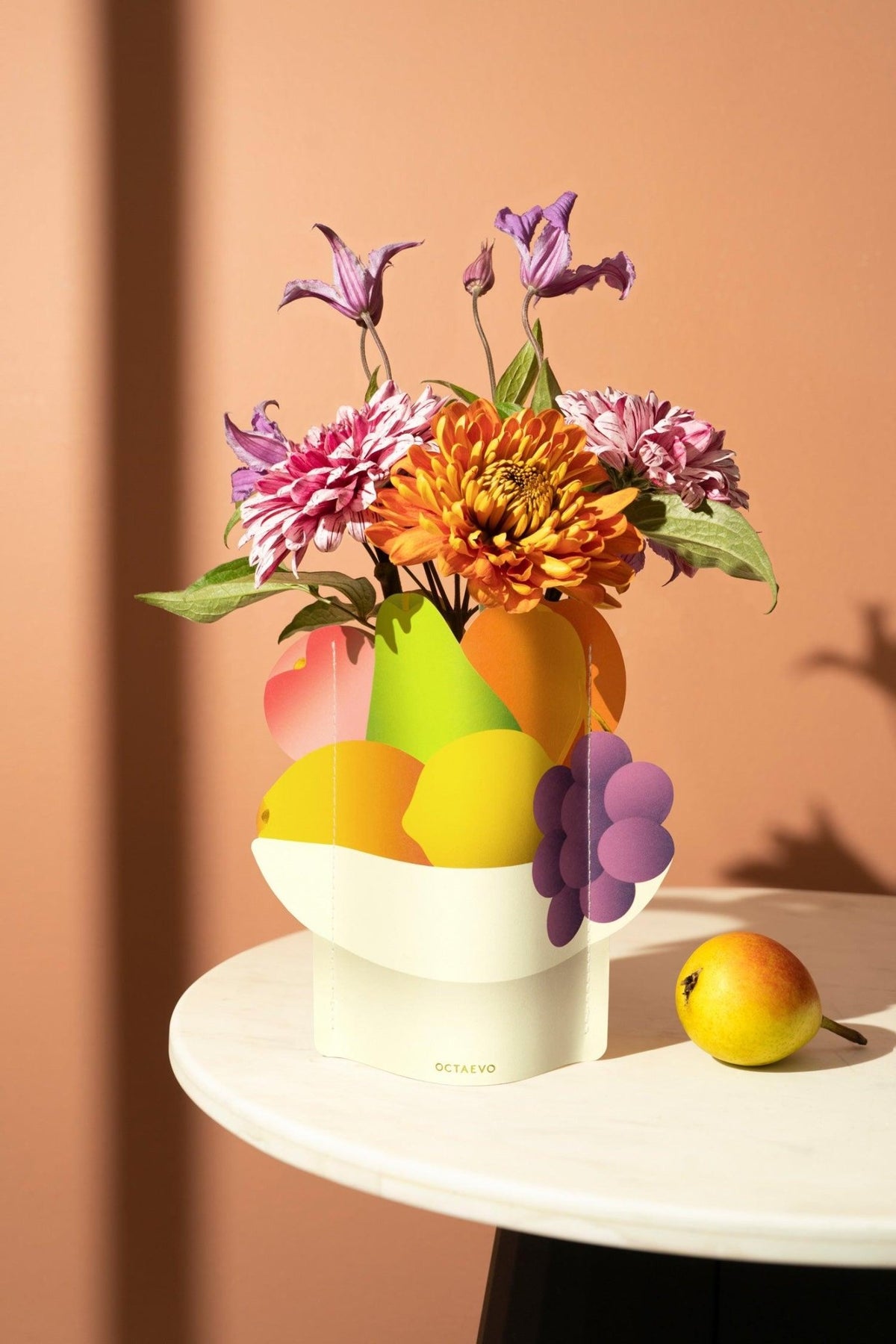 The Octaevo Fruit Bowl Mini Paper Vase with vibrant flowers sits on a white table against a peach-colored wall.