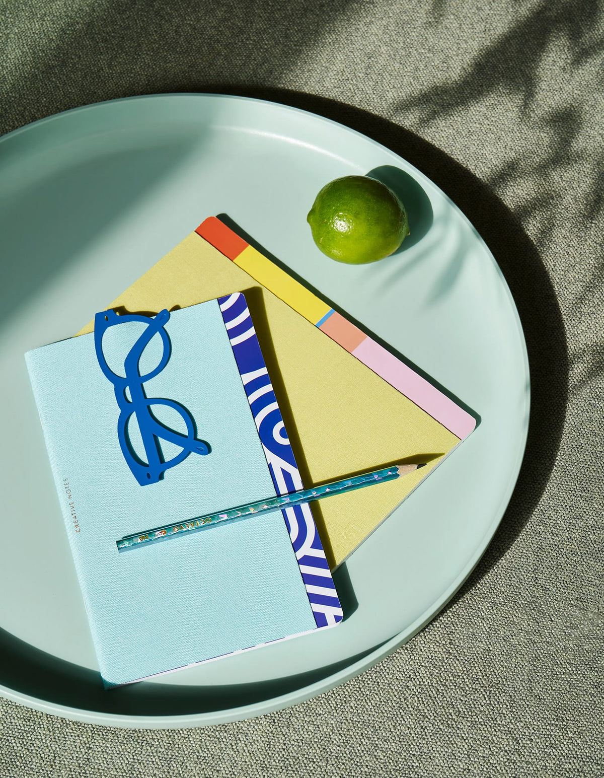 A lime, three notebooks, blue glasses, and a pencil on a large light-blue tray in sunlight.