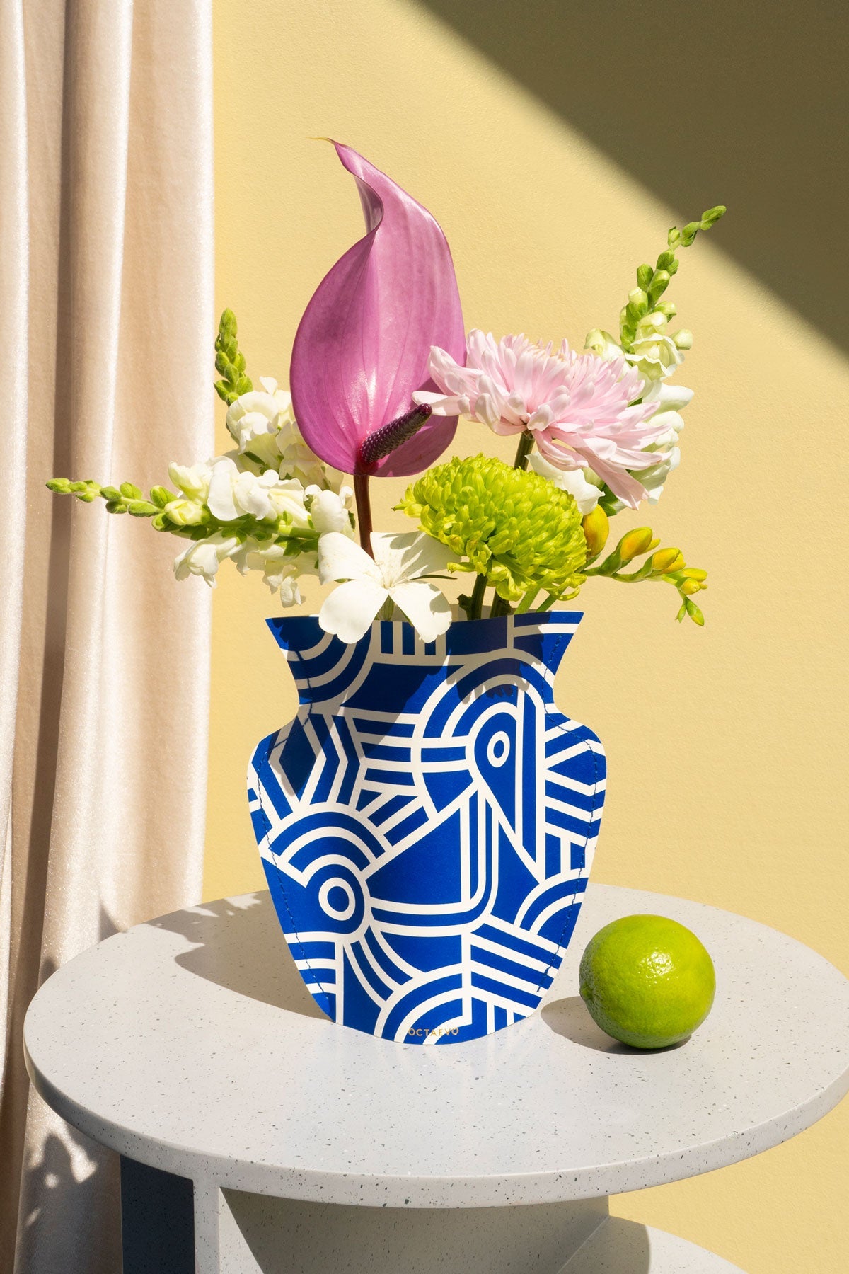 The Octaevo Greco Mini Paper Vase with blue and white patterns holds flowers on a table beside a green lime.