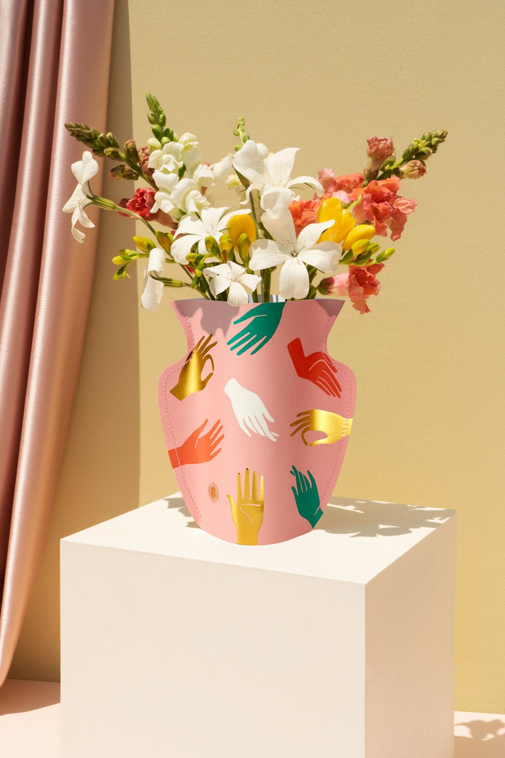 A pink OCTAEVO paper vase decorated with colorful hand illustrations, filled with flowers, displayed on a pedestal.