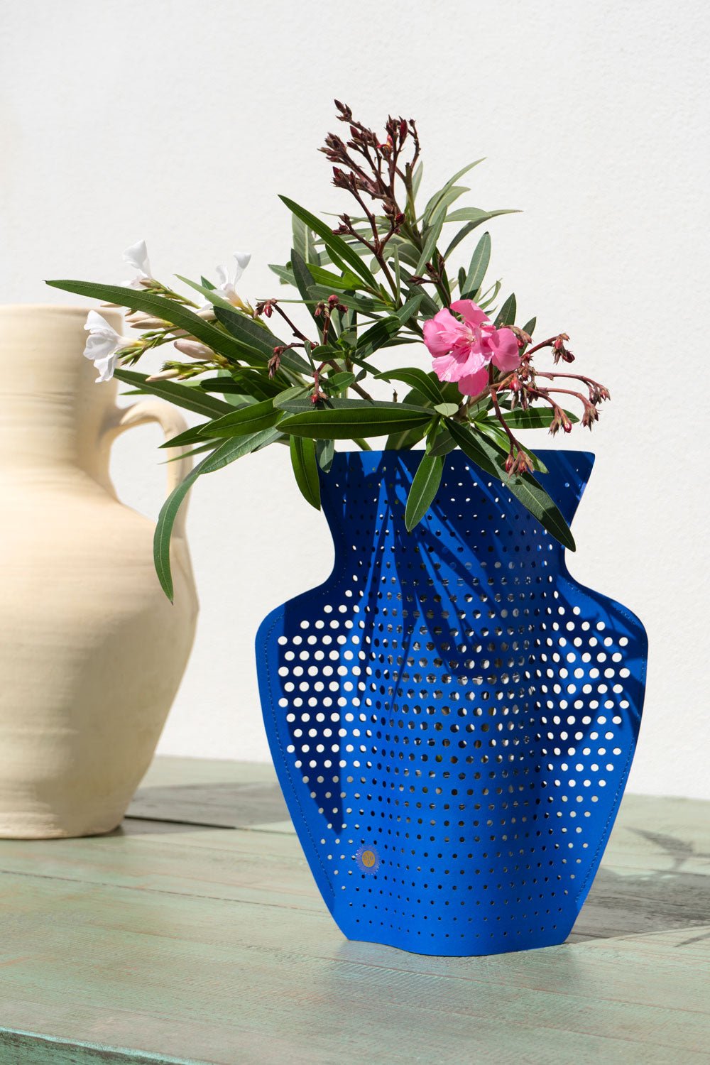 The Helio Paper Vase by OCTAEVO with blooms sits on a green table beside a beige jug against a white wall.
