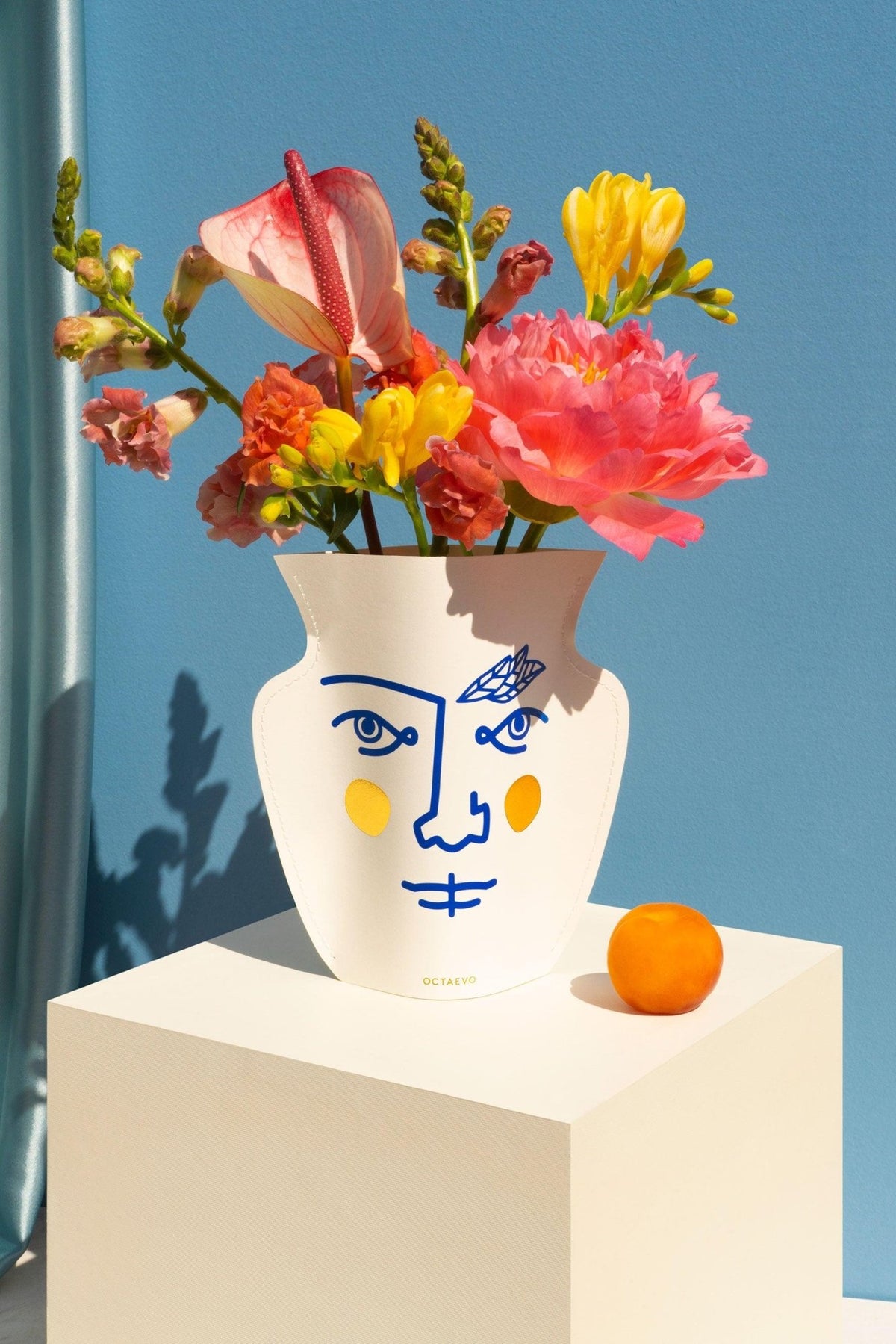 The OCTAEVO Janus Mini Paper Vase with a blue face holds flowers next to an orange on a white pedestal.
