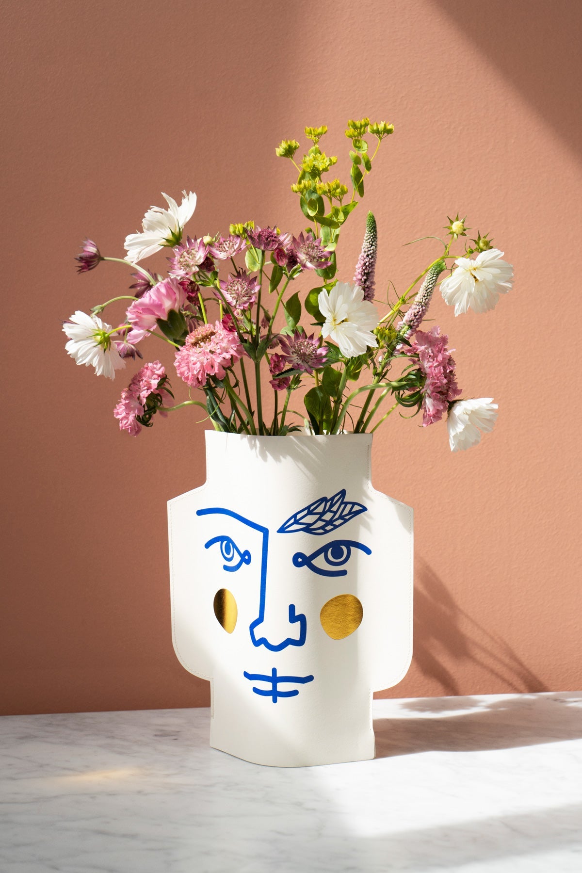 The OCTAEVO Janus Paper Vase features a blue abstract face and holds pink flowers on marble against a pink wall.