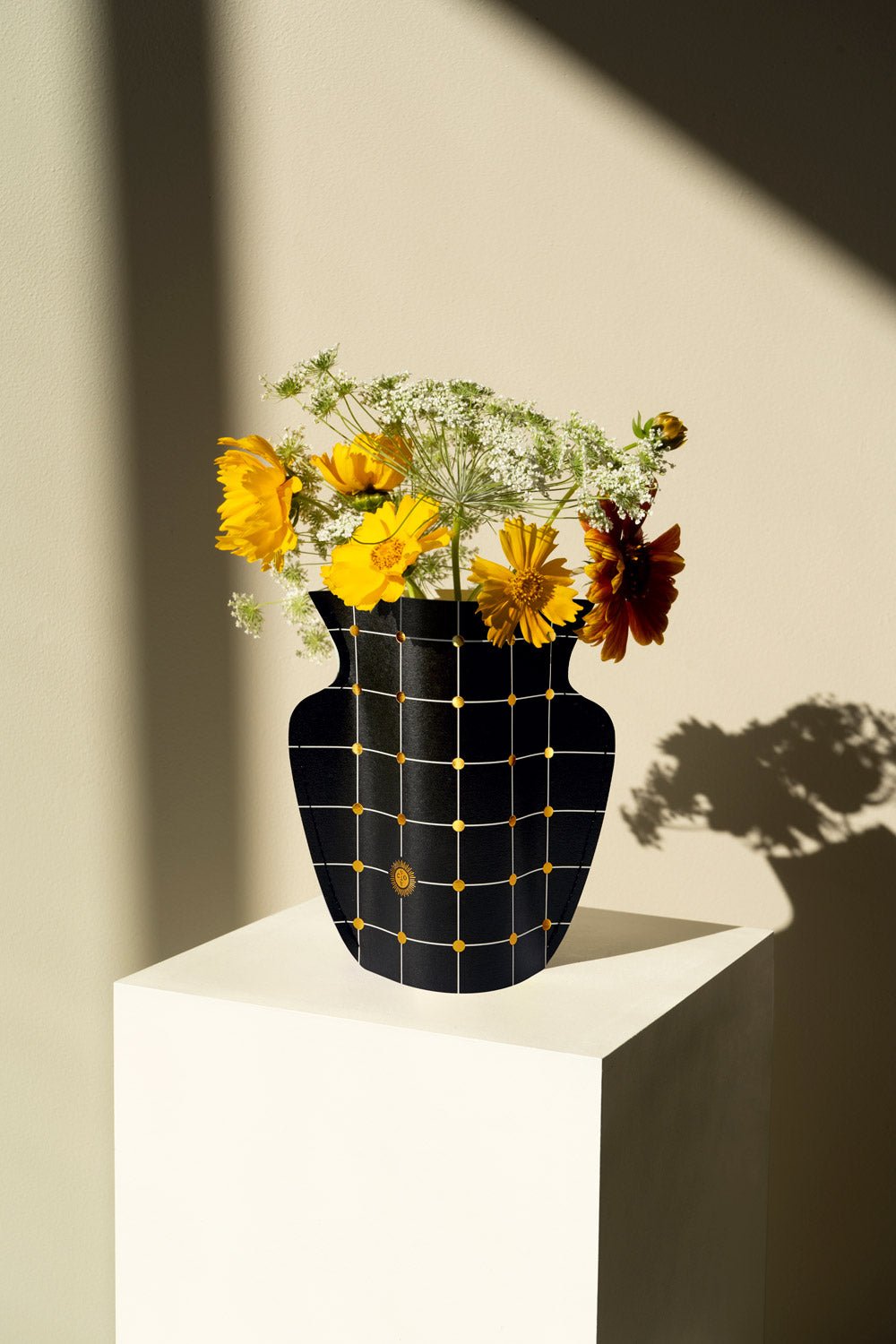 The Lido Mini Paper Vase by OCTAEVO features a yellow grid and casts an elegant shadow on a sunlit pedestal.