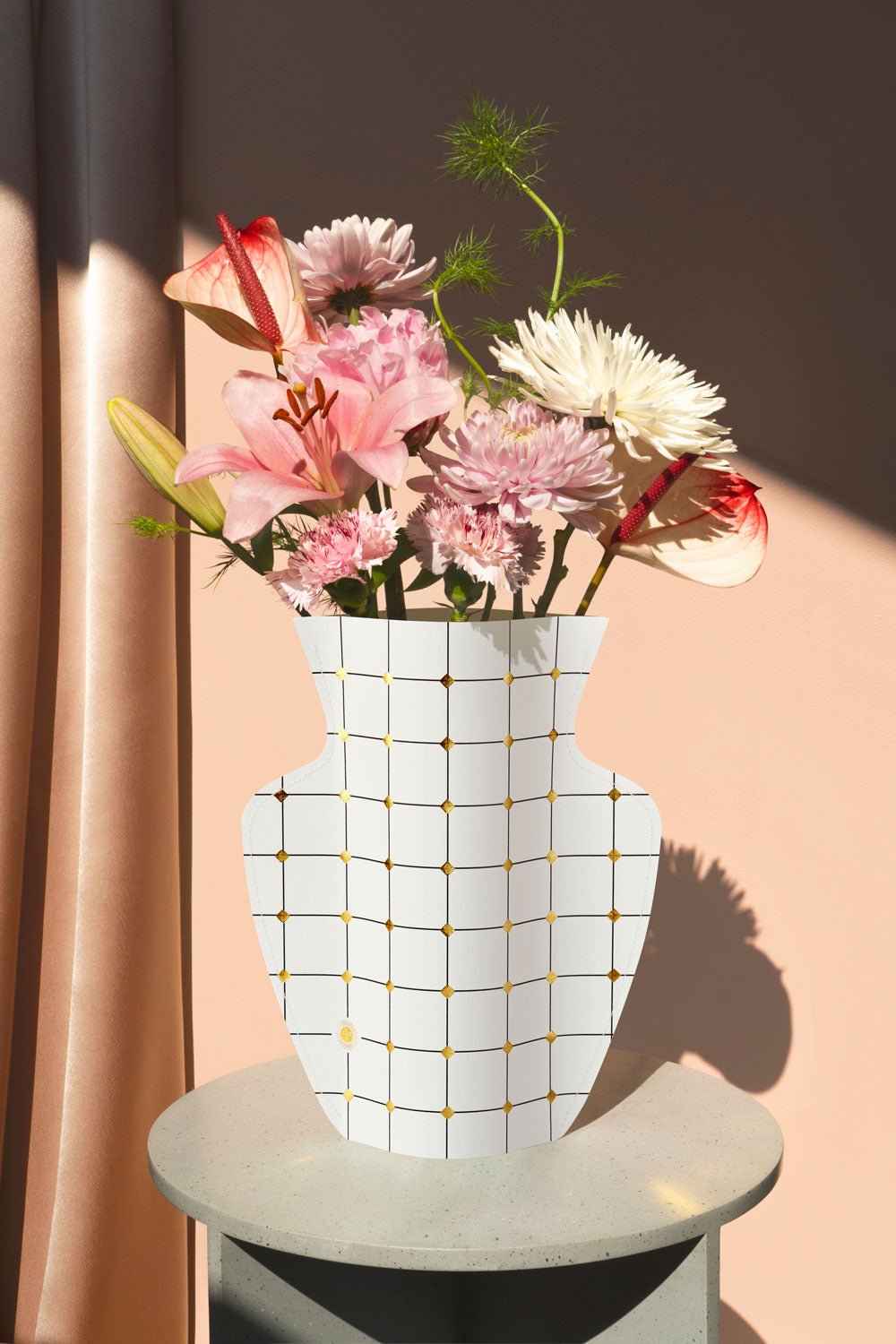 The OCTAEVO Lido Paper Vase with a grid design displays pink and white flowers on a sunlit round table.