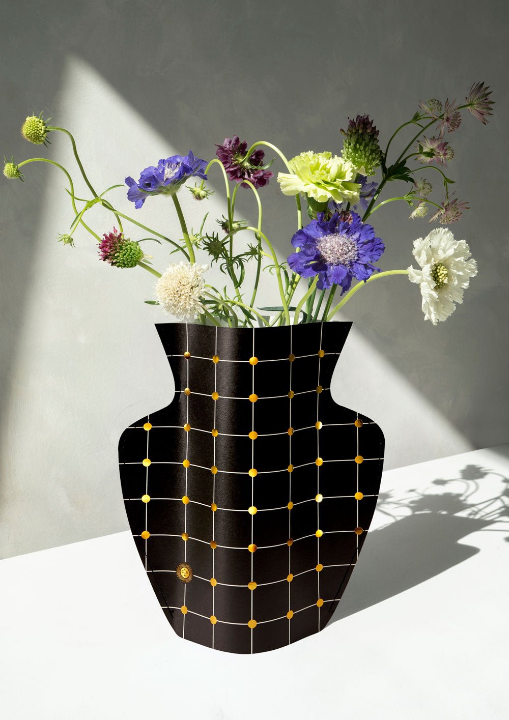 The OCTAEVO Lido Paper Vase with a black grid pattern displays wildflowers on a sunlit white surface.