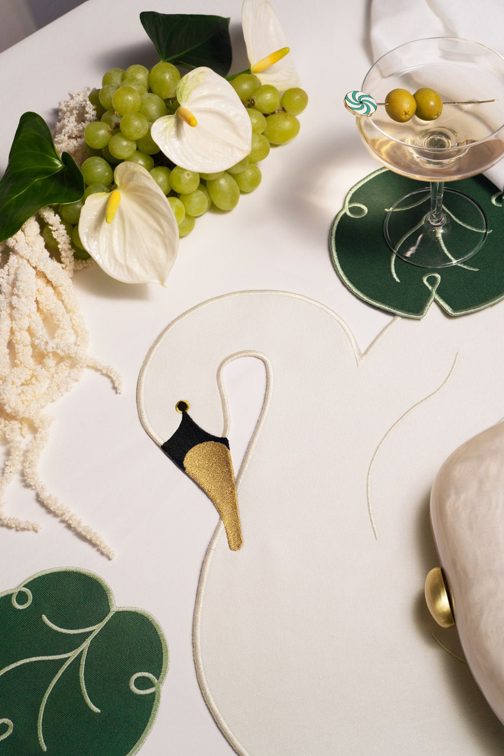 Une nappe blanche avec des cygnes, du raisin, des fleurs blanches et des dessous de verre Feuille de lys OCTAEVO, pour une table fraîche et élégante.