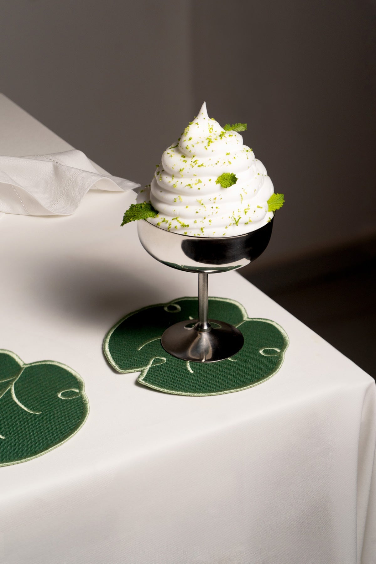 Une coupe en métal contenant un dessert à la chantilly repose sur des dessous de verre Feuille de lys OCTAEVO (lot de 4), sur une nappe blanche.