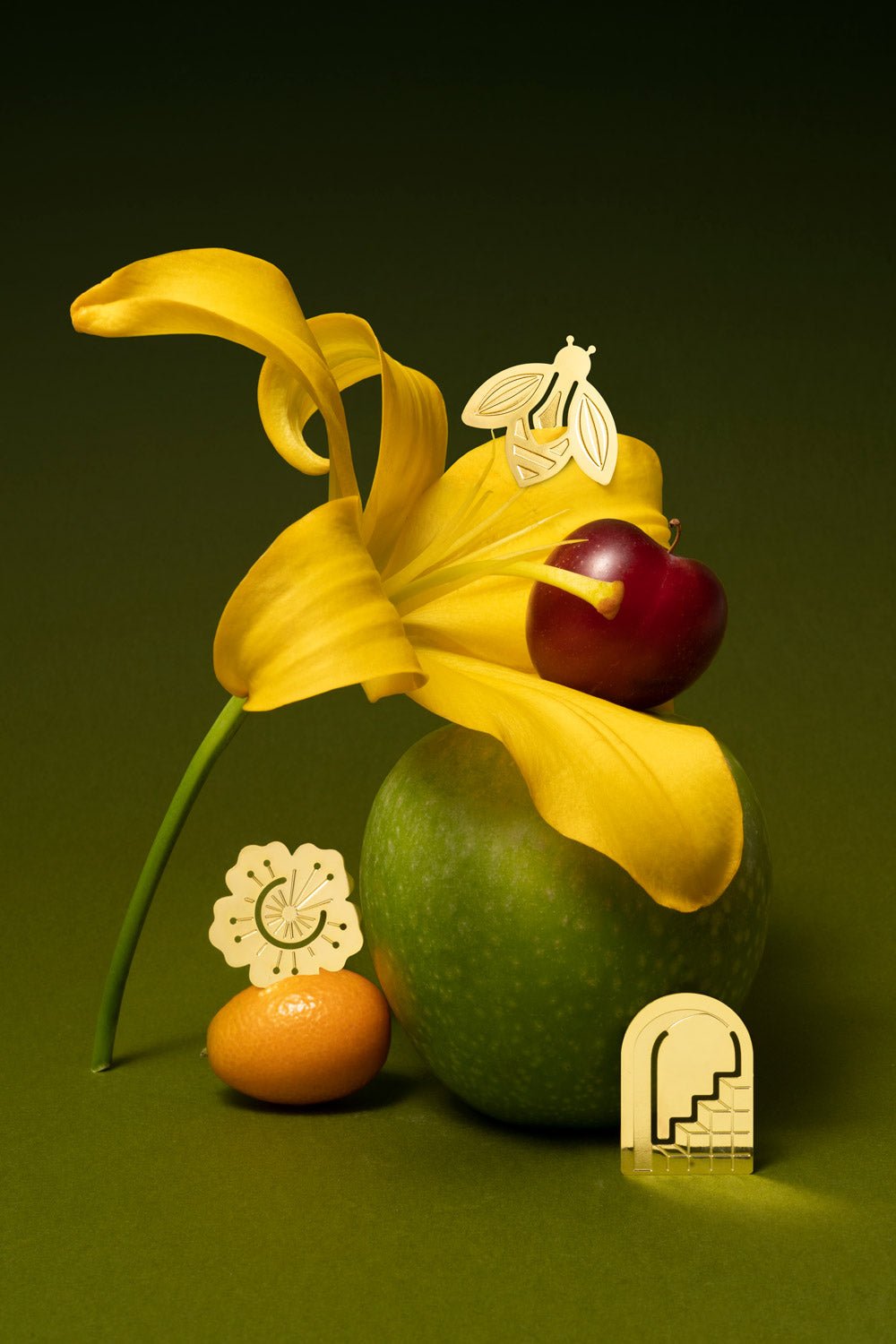 Three OCTAEVO Metal Clips Capri rest on a green background with a lily, green apple, kumquat, and cherry.