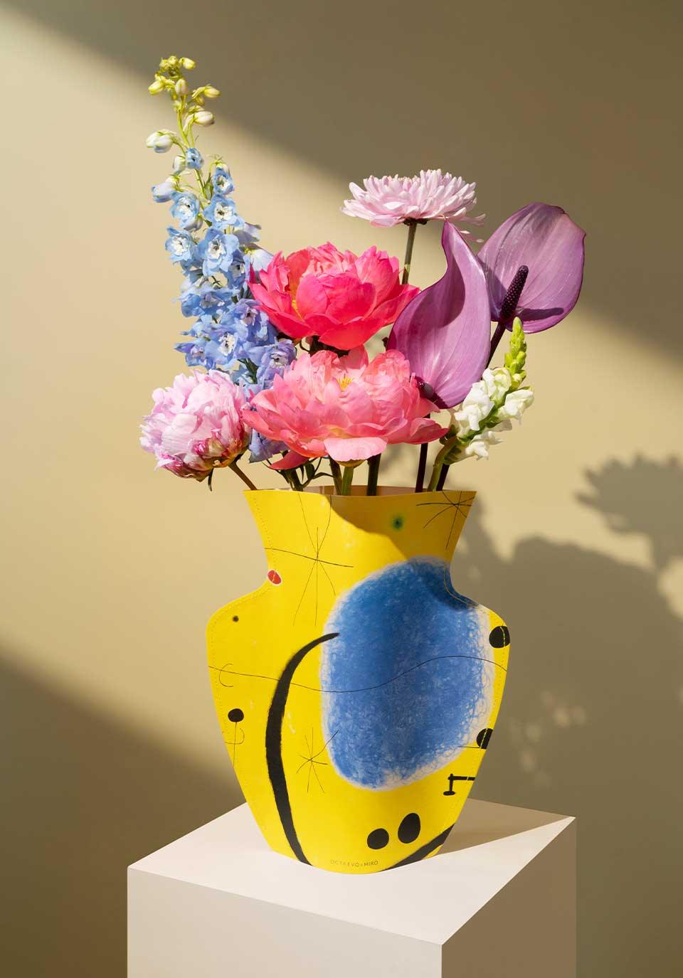 A bright yellow Octaevo Miró Paper Vase with abstract designs holds pink, purple, and blue flowers on a white pedestal.
