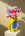 A bright yellow Octaevo Miró Paper Vase with abstract designs holds pink, purple, and blue flowers on a white pedestal.