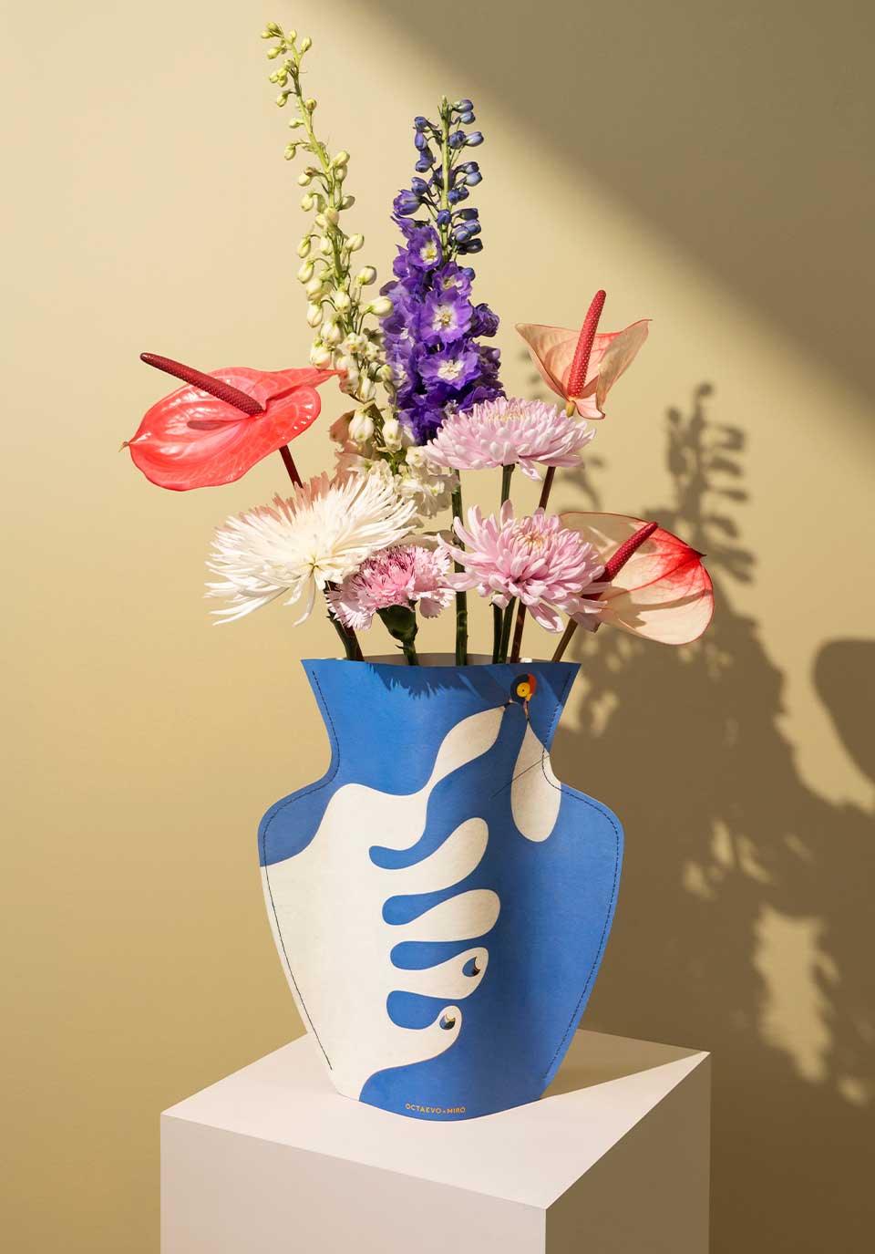 The Miró Paper Vase by Octaevo features a colorful bouquet in a white abstract hand, shown on a pedestal against beige.
