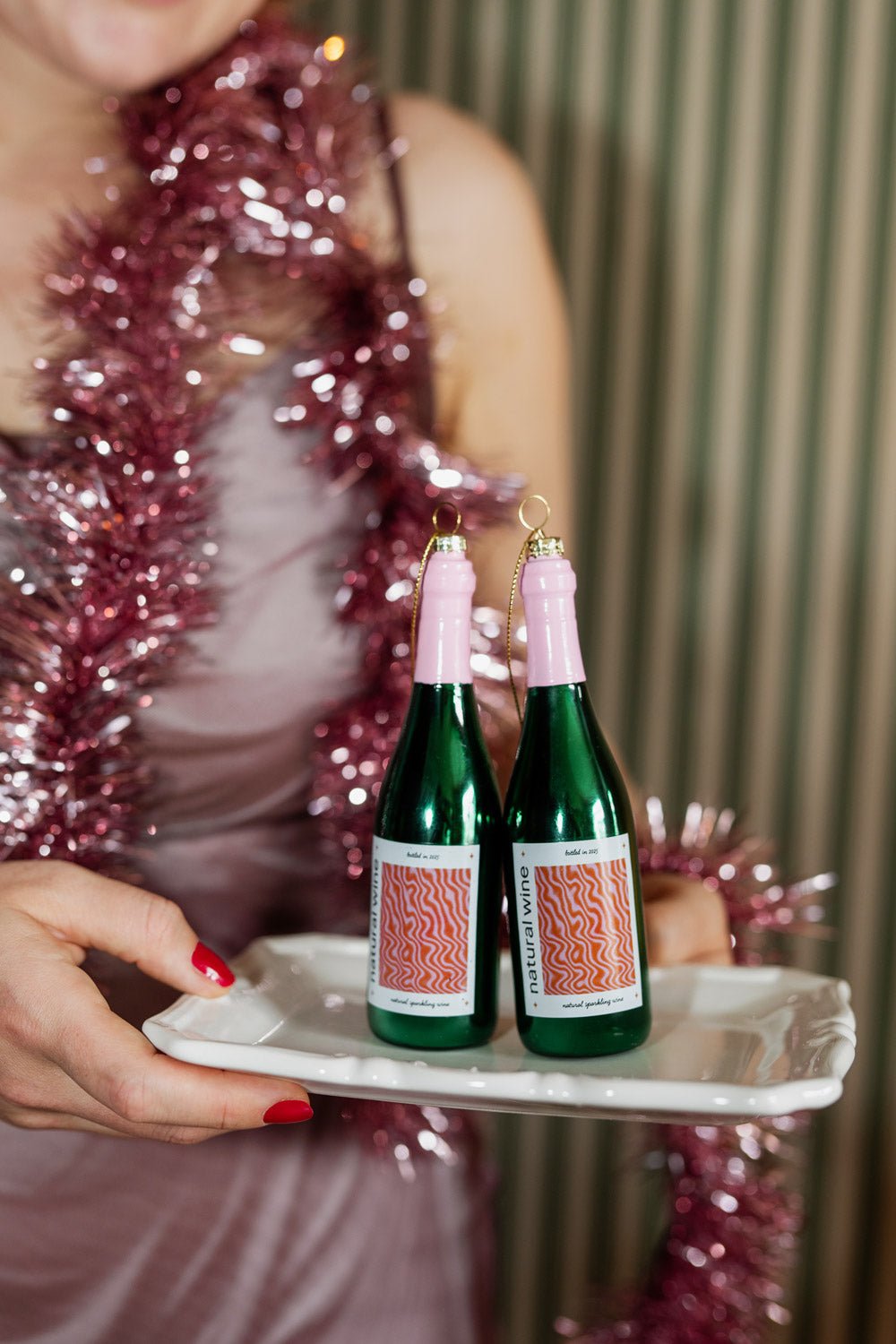 Person in a pink dress with tinsel holds a white tray with two OCTAEVO Natural Wine Ornaments.