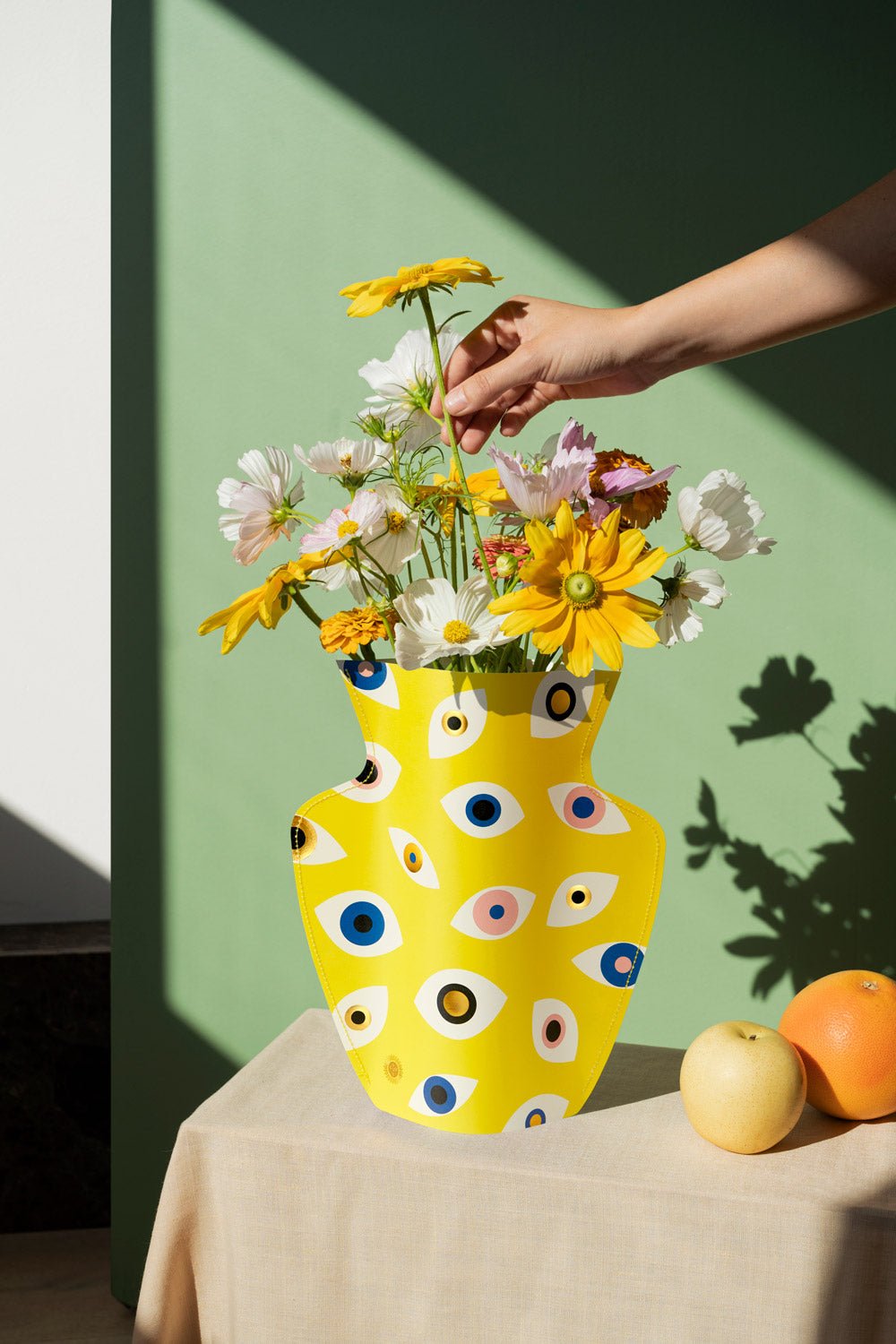 A hand arranges flowers in the OCTAEVO Nazar Paper Vase with eye motifs, on a sunny table with oranges.